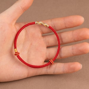 May include: A red braided bracelet with gold-colored accents. The bracelet features a twisted gold-colored center section and small gold-colored details. The bracelet is shown on a person's hand.