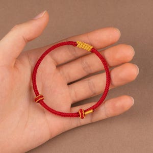 May include: A red braided bracelet with gold-colored accents. The bracelet features a simple, circular design with decorative knots and a gold clasp. The bracelet is shown against a neutral background.