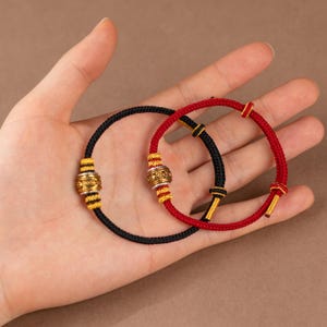 May include: Two handmade bracelets. One is black with gold and yellow accents, the other is red with black and gold accents. Both bracelets have a central decorative bead and are made of woven cord.