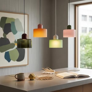 Op de afbeelding: Vier hanglampen op een rij, elk met een ander kleurenschema. De lampen hebben een cilindrische basis en een rechthoekige kap. Kleuren zijn groen, oranje, groen en roze. De lampen hangen boven een aanrecht met een mok, boek en decoratieve items.