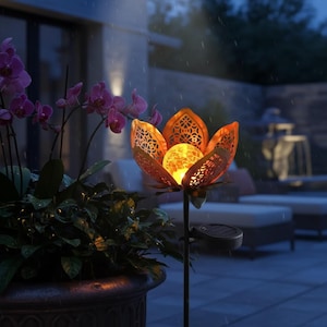 Könnte beinhalten: Eine solarbetriebene Gartenleuchte in Form einer Lotusblume. Die orangefarbenen Metallblätter haben ein dekoratives Ausschnittmuster, das eine leuchtende Glaskugel umschließt. Der Pfahl befindet sich in einem Garten mit lila Orchideen und einem dunklen Hintergrund.