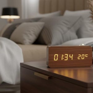 Puede incluir: Un reloj despertador digital de madera que muestra la hora 1:34 y 20°C está sobre una mesita de noche. El reloj tiene forma rectangular con acabado de vetas de madera. Un jarrón negro, un jarrón blanco y ramitas decorativas también están sobre la mesa.