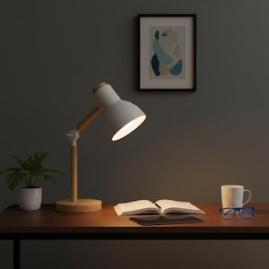 May include: A white desk lamp with a wooden arm and base illuminates a desk. An open book, a tablet, a mug, and glasses sit on the wooden desk. A small potted plant and a framed abstract art piece are also visible.