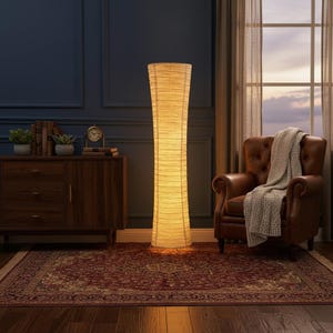 May include: A tall, beige floor lamp with a textured, cylindrical shade illuminates a room. The lamp stands next to a brown leather armchair with a grey blanket. A dark wooden cabinet and a patterned rug are also visible.