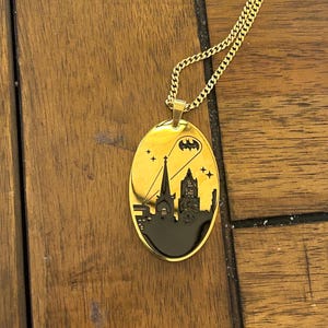 May include: Gold-colored oval pendant necklace featuring a black cityscape silhouette with a Batman symbol and stars. The pendant hangs from a matching gold chain. The necklace is displayed on a wooden surface.