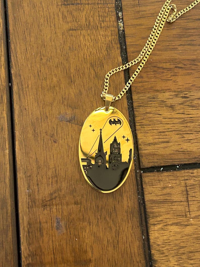 May include: Gold-colored oval pendant necklace featuring a black cityscape silhouette with a Batman symbol and stars. The pendant hangs from a matching gold chain. The necklace is displayed on a wooden surface.