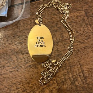 Peut inclure: Collier dor&eacute; avec un pendentif ovale portant l'inscription "THIS IS A LOVE STORY" en noir. La cha&icirc;ne est de type maillon. Le collier est pr&eacute;sent&eacute; sur une surface en bois.