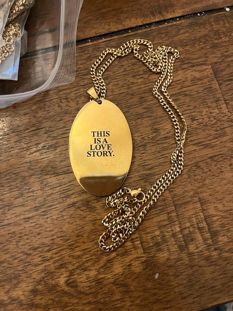 Peut inclure: Collier dor&eacute; avec un pendentif ovale portant l'inscription "THIS IS A LOVE STORY" en noir. La cha&icirc;ne est de type maillon. Le collier est pr&eacute;sent&eacute; sur une surface en bois.