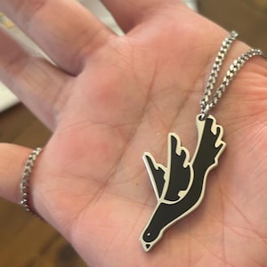 May include: A silver-toned metal pendant shaped like a bird in flight, featuring a black enamel inlay on the wings and body. The pendant is attached to a silver-toned metal curb chain necklace with small, uniform links that drape across the palm.