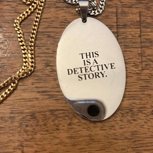 May include: A silver-colored pendant necklace with the text "THIS IS A DETECTIVE STORY." The pendant is oval-shaped and hangs from a silver chain. A gold chain is also visible in the image.