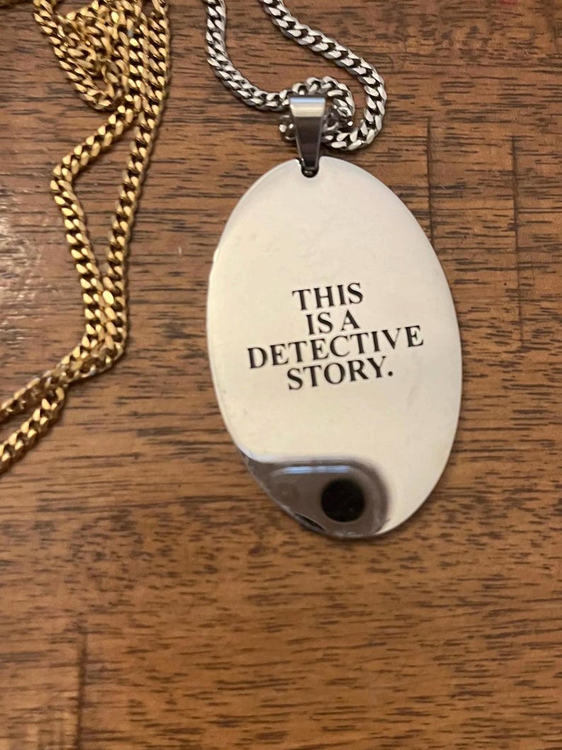 May include: A silver-colored pendant necklace with the text "THIS IS A DETECTIVE STORY." The pendant is oval-shaped and hangs from a silver chain. A gold chain is also visible in the image.