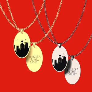 Peut inclure: Quatre colliers avec des pendentifs ovales. Deux sont dor&eacute;s, deux sont argent&eacute;s. Chaque pendentif pr&eacute;sente une silhouette de paysage urbain noir et le texte "THIS IS A LOVE STORY". Le fond est rouge uni.