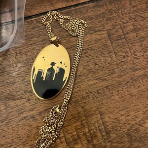 Peut inclure: Collier dor&eacute; avec un pendentif ovale repr&eacute;sentant une silhouette de paysage urbain noir. Le pendentif a un fond dor&eacute; avec un motif de skyline noir et une cha&icirc;ne dor&eacute;e.