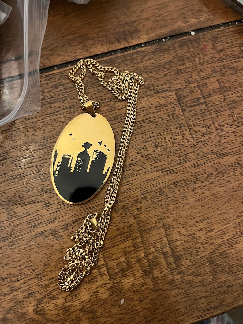 Peut inclure: Collier dor&eacute; avec un pendentif ovale repr&eacute;sentant une silhouette de paysage urbain noir. Le pendentif a un fond dor&eacute; avec un motif de skyline noir et une cha&icirc;ne dor&eacute;e.