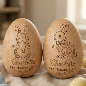 May include: Two wooden Easter egg ornaments. Each egg features a laser-etched bunny design, the name "Charlotte", and the text "First Easter 2026". One bunny holds an egg with "1st" on it. The eggs are light brown.
