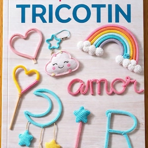 May include: A book cover with the title "Moldes para crear en TRICOTIN" in pink and blue. The image features various crochet items, including a rainbow, hearts, stars, a cloud, and the word "amor".