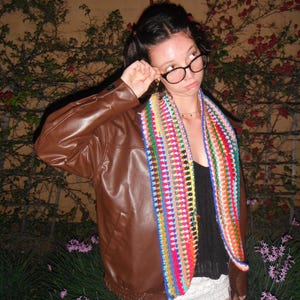 May include: A brown leather jacket and a colorful crochet scarf are worn over a black top and white shorts. The scarf features a vibrant array of colors, including red, blue, green, and yellow. The outfit is worn outdoors.