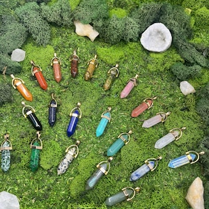 Peut inclure: Une collection de pendentifs en pierres précieuses de différentes couleurs, notamment orange, bleu, vert et rose, avec des accents métalliques dorés. Les pendentifs sont présentés sur un lit de mousse verte, avec une géode blanche et des cristaux de quartz en arrière-plan.