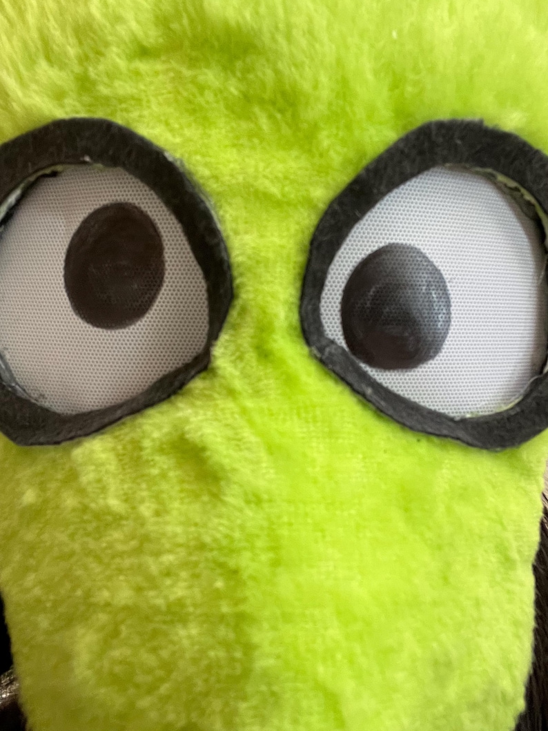 May include: Close-up of a plush, lime green monster face with large, round eyes. The eyes have white mesh pupils with dark brown centers, framed by black felt. The fur is soft and textured.