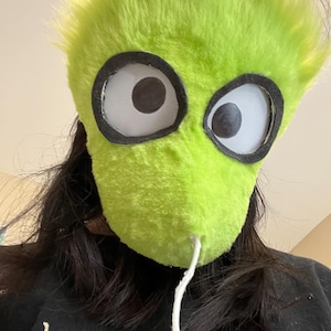 May include: A lime green, furry mask with large, cartoonish eyes. The mask has a white string attached to the bottom. The person wearing the mask is wearing a black shirt with the words "hear vibes..." printed on it.