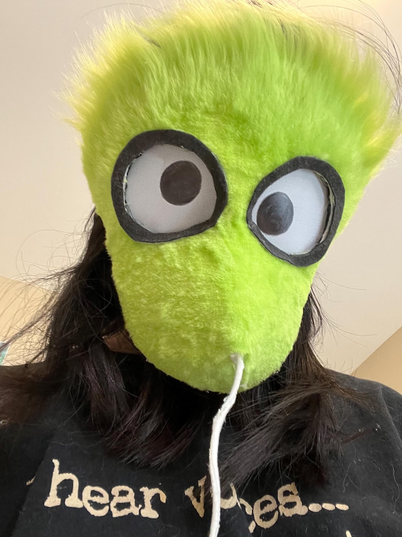 May include: A lime green, furry mask with large, cartoonish eyes. The mask has a white string attached to the bottom. The person wearing the mask is wearing a black shirt with the words "hear vibes..." printed on it.