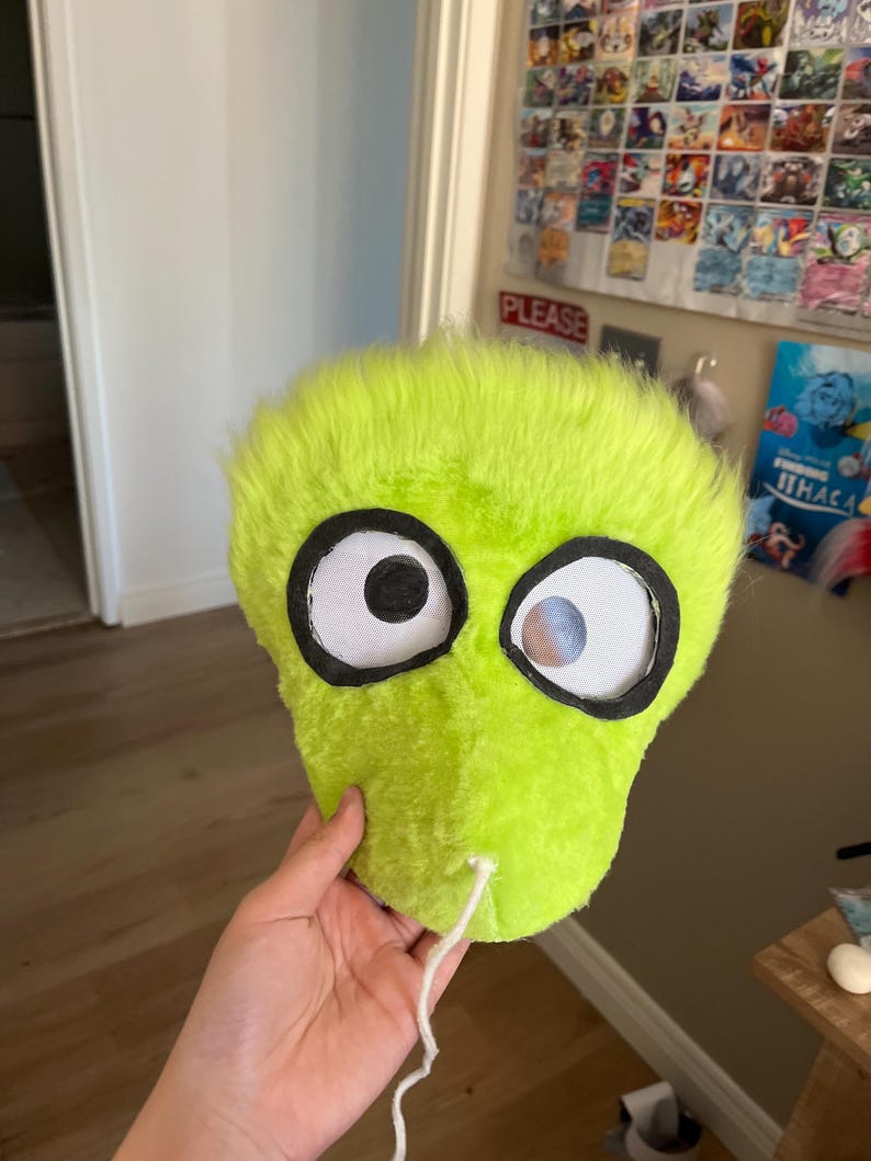 May include: A lime green plush mask with large, cartoonish eyes outlined in black. The eyes have white centers with a small blue and gray detail. A white string extends from the bottom of the mask.
