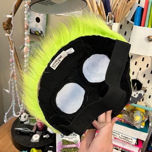 May include: A black and neon green furry mask with white eye openings and an elastic strap. The mask has a tag that reads "Stickbuggy Studios". The mask is likely for cosplay or costume use.