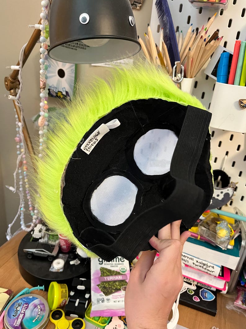 May include: A black and neon green furry mask with white eye openings and an elastic strap. The mask has a tag that reads "Stickbuggy Studios". The mask is likely for cosplay or costume use.