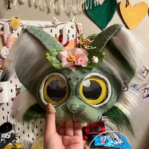 Può includere: Una maschera di creatura fantasy verde e bianca con grandi occhi gialli e accenti di pelliccia bianca. La maschera è decorata con fiori rosa e piccoli abbellimenti. La maschera è tenuta da una mano.