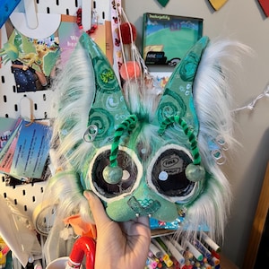 May include: A handmade, whimsical mask with large, expressive eyes and pointed ears. The mask features shades of green, white, and teal, with fluffy white faux fur accents. Decorative elements include green pipe cleaners and silver-colored rings.