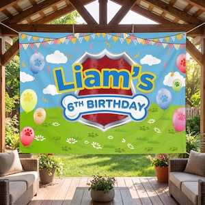 May include: A blue banner with the text "Liam's 6th Birthday" in yellow and white. The banner features a dog bone design, paw print balloons, and a green grass background. The banner is hanging outdoors.