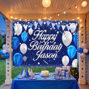 May include: A dark blue birthday backdrop featuring the silver text Happy Birthday Jason, decorated with blue and silver glitter balloons, star patterns, and blue bunting flags, displayed above a table with a cake and gift boxes.