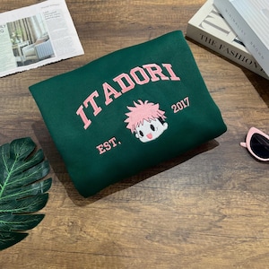 May include: A dark green sweatshirt with the word "ITADORI" in pink embroidered lettering. Below the word is a cartoon character with pink hair and the year "2017". The word "EST." is below the character.