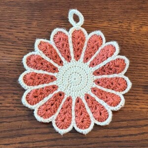 May include: A handmade crochet flower-shaped potholder. The petals are coral, edged with white, and surround a cream-coloured centre. A small loop is attached for hanging. The potholder is on a wooden surface.