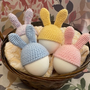 以下が含まれることがあります： 柳のバスケットには、4つのイースターエッグの装飾品が入っています。各卵には、青、ピンク、黄色、ラベンダーのパステルカラーの、かぎ針編みのウサギの帽子が被せられています。卵は黄色の布の上に置かれています。