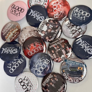 May include: A collection of circular pin-back buttons featuring various designs, including text such as Blood Saga, The X: Nexus, and Arirang MetLife Stadium. The buttons display diverse graphics, including photographic portraits, stadium views, and patterns.