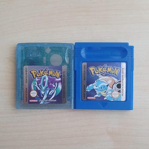 Può includere: Due cartucce di gioco Pokemon Game Boy vintage. Una è blu con una grafica di Blastoise, l'altra è blu chiaro con una grafica di Suicune. Entrambe hanno il logo Pokemon e testo in tedesco. Le cartucce sono su una superficie marrone chiaro.