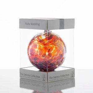 May include: A spherical glass ornament with a swirling pattern of deep red, orange, and white colors. It is housed in a clear square display box labeled Ruby Wedding and The Sienna Collection, featuring text that reads Inspirational Hand Crafted Art Glass.