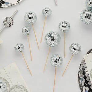 Op de afbeelding: Een verzameling zilveren discobal taarttoppers op houten stokjes. De spiegelbollen variëren in grootte en zijn gerangschikt op een wit oppervlak, wat wijst op een feest- of thema-evenement. Extra discobaldecoraties zijn ook aanwezig.