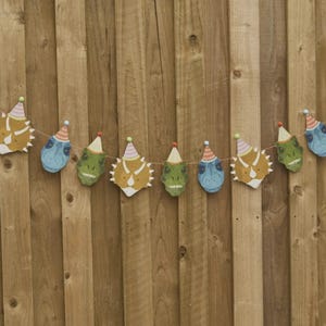 May include: A string of dinosaur head cutouts in shades of blue, green, and yellow, each wearing a party hat. The garland is strung on a thin brown string and hangs against a wooden fence background.