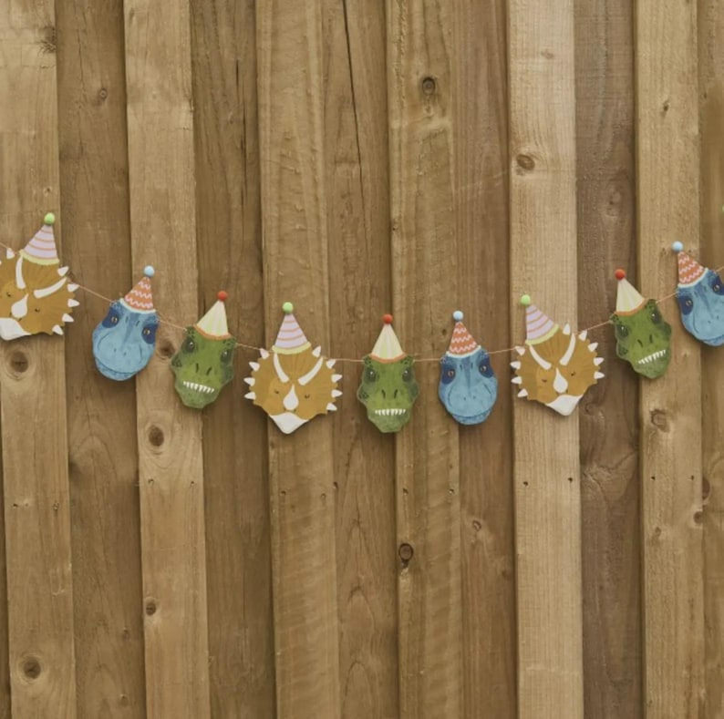May include: A string of dinosaur head cutouts in shades of blue, green, and yellow, each wearing a party hat. The garland is strung on a thin brown string and hangs against a wooden fence background.