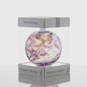 Puede incluir: Una esfera decorativa de vidrio con tonos morados, rosas y dorados, presentada en una vitrina transparente. La parte superior de la caja dice "Pearl Wedding". La base de la caja tiene el texto "The Sienna Collection".