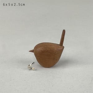 Puede incluir: Una pequeña figura de pájaro de madera marrón con un cuerpo liso y redondeado, un pico puntiagudo y una cola erguida. El objeto mide 6 x 5 x 2,5 cm. Una pequeña ramita de flores secas blancas está colocada cerca de la base de la figura.