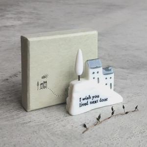 Puede incluir: Una figura de casa de cerámica blanca con tejados azules y un árbol, junto a una caja verde claro. La figura tiene el texto "I wish you lived next door". Una ramita de capullos está en primer plano.
