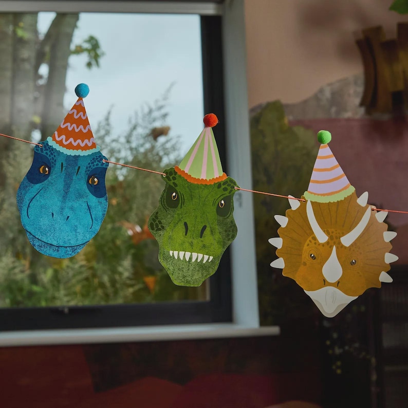May include: A string of dinosaur-shaped party decorations. The decorations feature a blue T-Rex, a green T-Rex, and a Triceratops, each wearing a party hat. The string is orange, and the background includes a window and a wall.