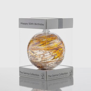 May include: A decorative glass orb with a swirling pattern of gold and amber hues, presented in a clear display box. The box is labeled "Happy 50th Birthday" and "The Sienna Collection."