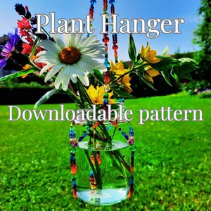 May include: A clear glass jar suspended by a colorful beaded plant hanger, containing a bouquet of white daisies, yellow flowers, and purple sprigs. The text Plant Hanger Downloadable pattern is overlaid on the image against a green lawn background.