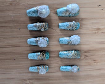 Op de afbeelding: Een set van tien kunstnagels met een turquoise en witte marmerafwerking en gouden metallic lijnen. Sommige nagels zijn versierd met witte bloemdecoraties en kleine witte parels, gerangschikt in twee verticale kolommen van vijf stuks.