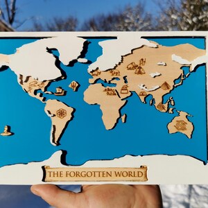 May include: A wooden world map with a blue background and a white frame. The map features laser-cut continents in a natural wood tone, with small icons representing landmarks. The text "THE FORGOTTEN WORLD" is at the bottom.