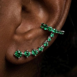 Op de afbeelding: Oor manchet oorbellen met een reeks heldergroene edelstenen. Het gebogen ontwerp volgt de contouren van het oor, met een rechte staaf die zich over de bovenkant van het oor uitstrekt. De edelstenen zijn gezet in een zilverkleurig metaal.